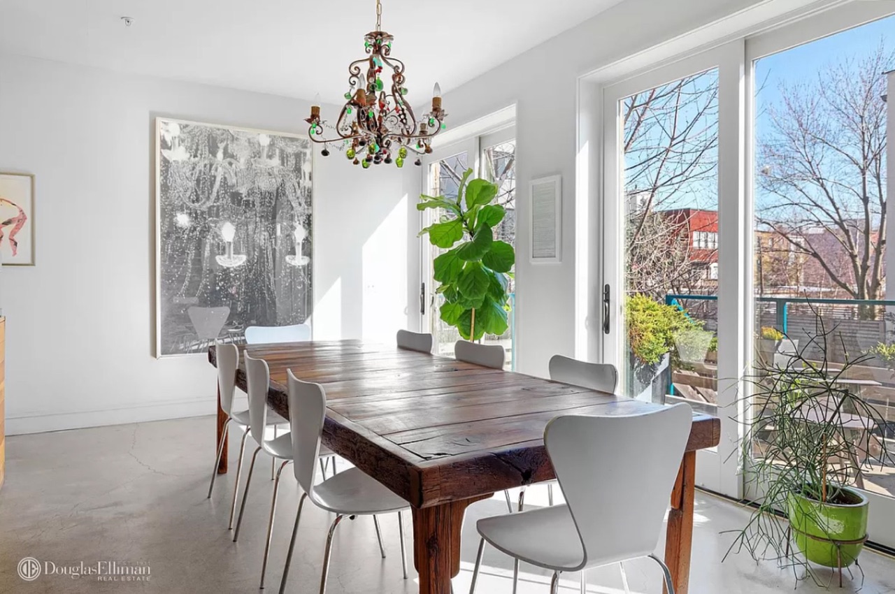 The dining room — live-edge dining table, antique-style chandelier overhead, large-scale abstract art against the white wall, floor-to-ceiling glass doors to the garden, fiddle-leaf fig and polished concrete