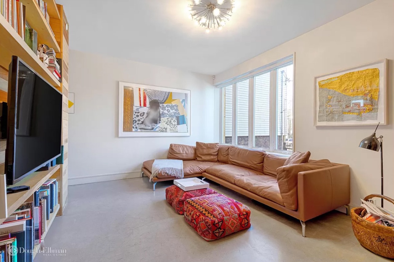 The living room — tan leather mid-century sectional, kilim floor cushion, the wood-clad bookshelf wall on the left with the TV mounted on a poplar panel, picture window above the sectional, polished concrete flooring