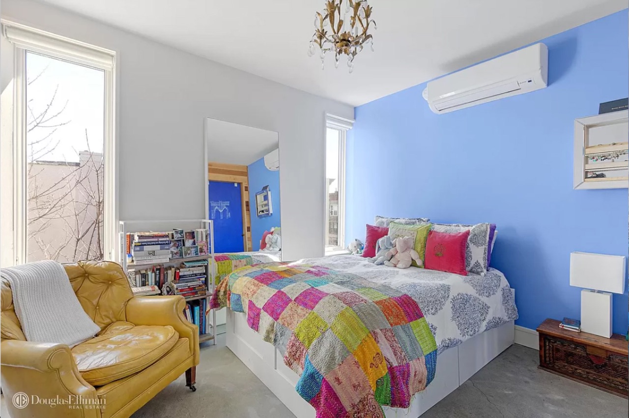 A secondary bedroom — vibrant blue accent wall, patchwork quilt, yellow leather armchair, white-painted vanity mirror, floor-to-ceiling windows