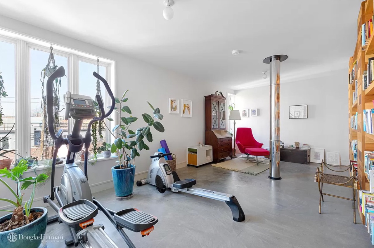 The upper-level gym / multi-use room — open space with elliptical and rowing machines, antique secretary desk, red modern lounge chair, bookshelves, floor-to-ceiling windows opening to the roof deck beyond