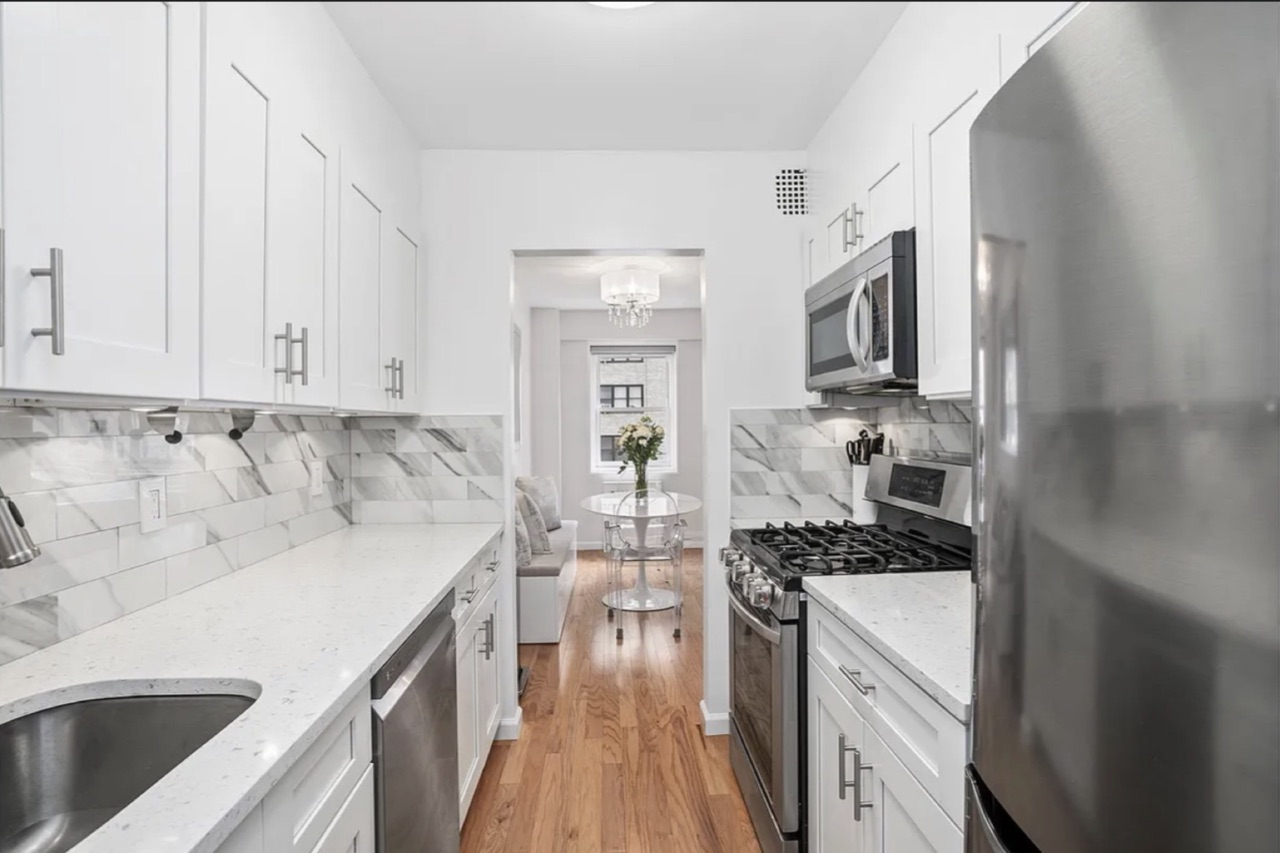 The renovated galley kitchen — white shaker cabinetry, marble herringbone backsplash, quartz countertops, stainless appliances, with sightlines through to the dining alcove at the rear