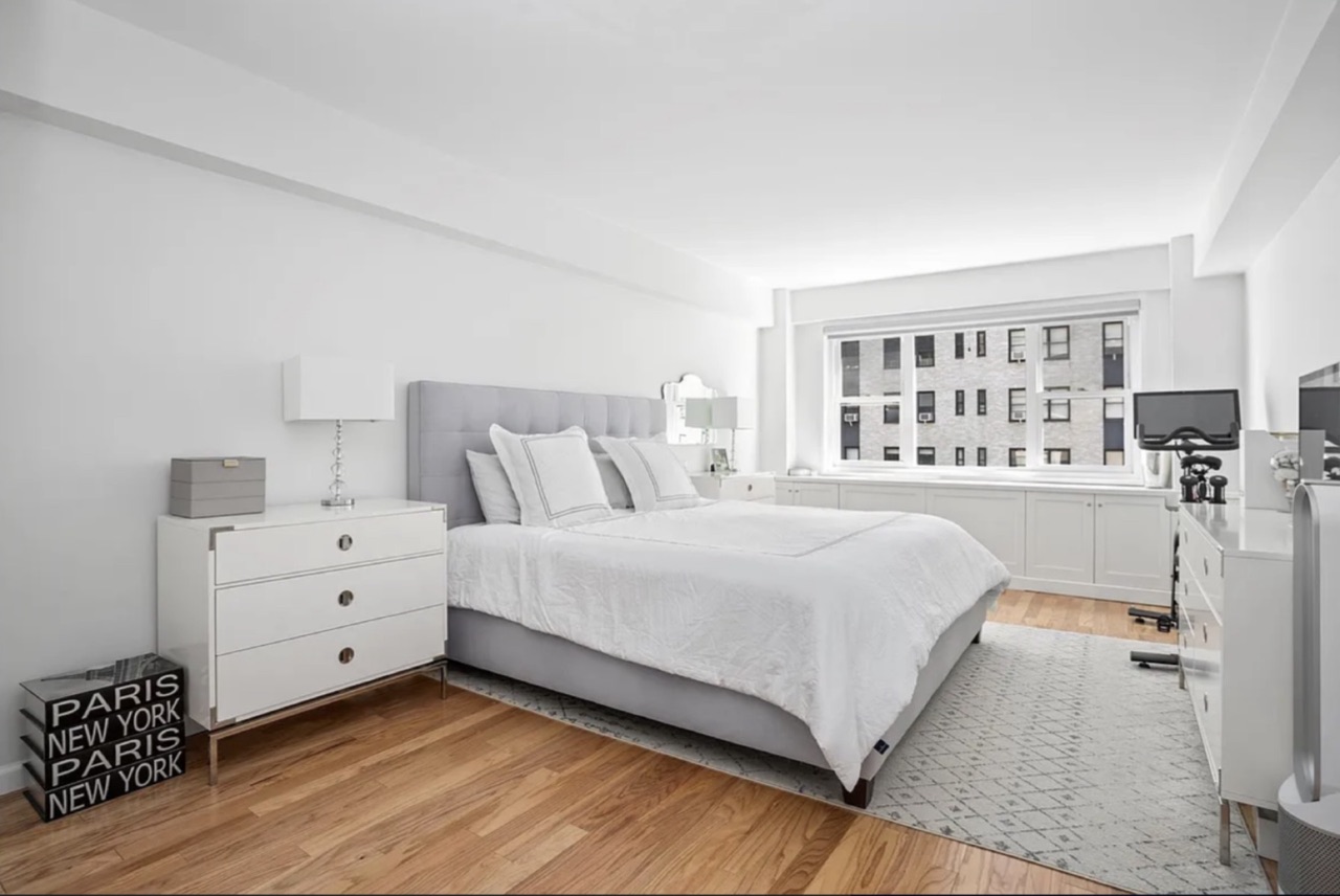 The primary bedroom — grey upholstered headboard, window over building-courtyard exposure, substantive room-scale and built-in storage along the window wall