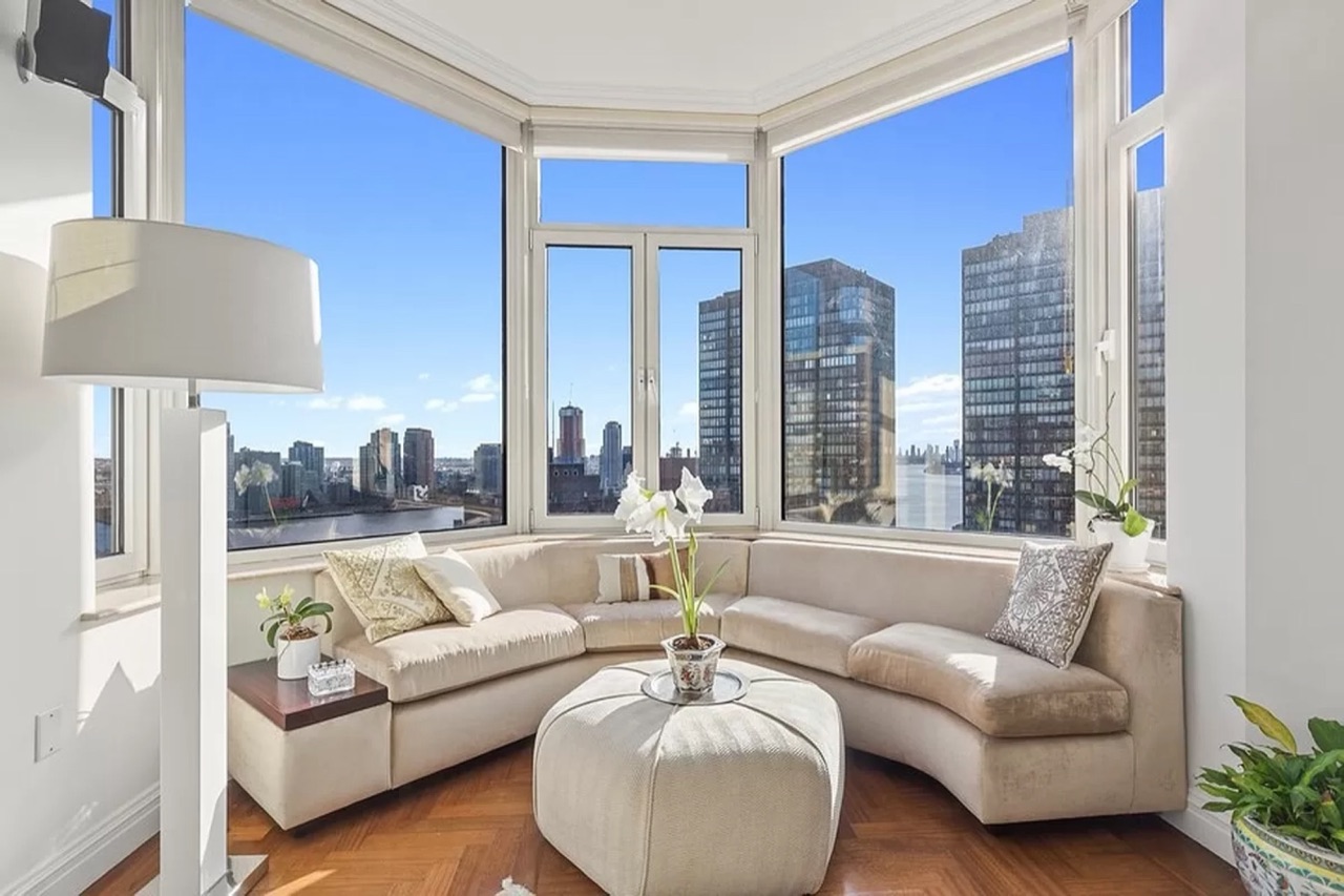 Grand Beekman #21C — the bay-windowed living room with panoramic East River and Long Island City skyline views, the defining architectural feature of the C-line