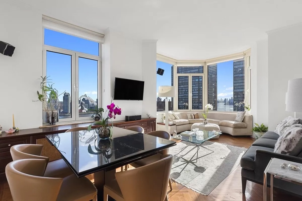 The open-plan living and dining flow — bay-window seating, dining table with leather chairs, dual exposure light across both the south windows and the east-facing river-view bay
