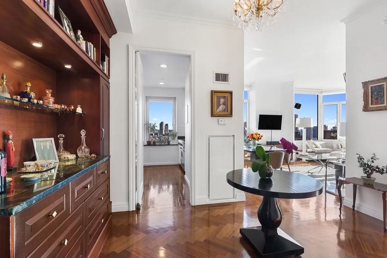 The entry foyer — built-in mahogany breakfront with green-marble counter, crystal chandelier overhead, sightlines through to the kitchen on the left and the living room on the right